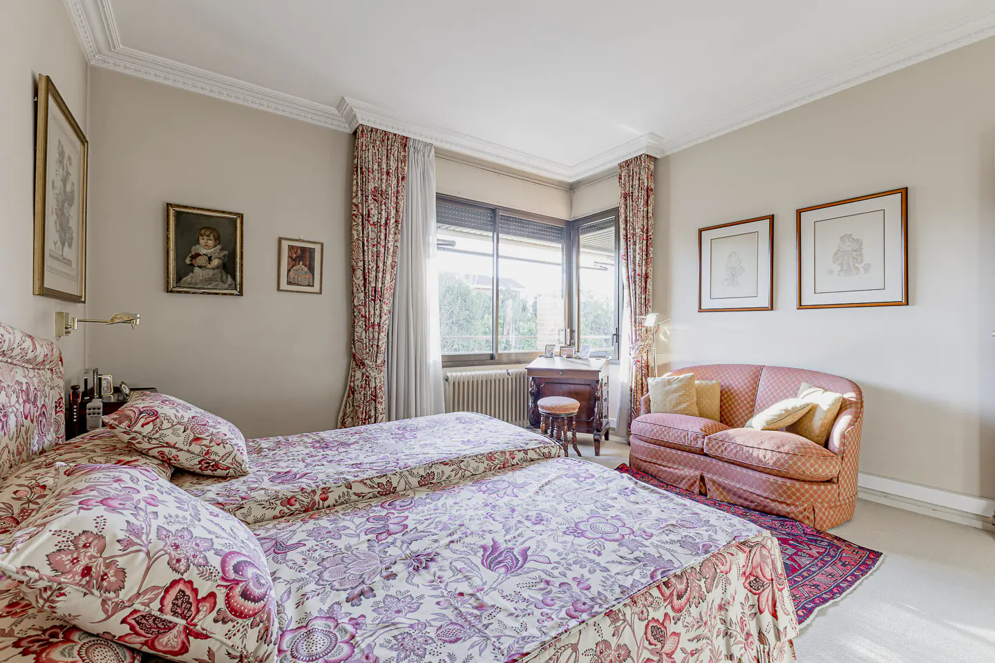 Bedroom with two twin beds, floral bedding, pink sofa, desk by window, and framed art on the walls.