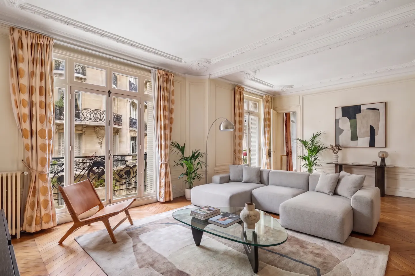 Bright living room with a gray sectional sofa, glass coffee table, and a tan leather chair near a window with patterned curtains.