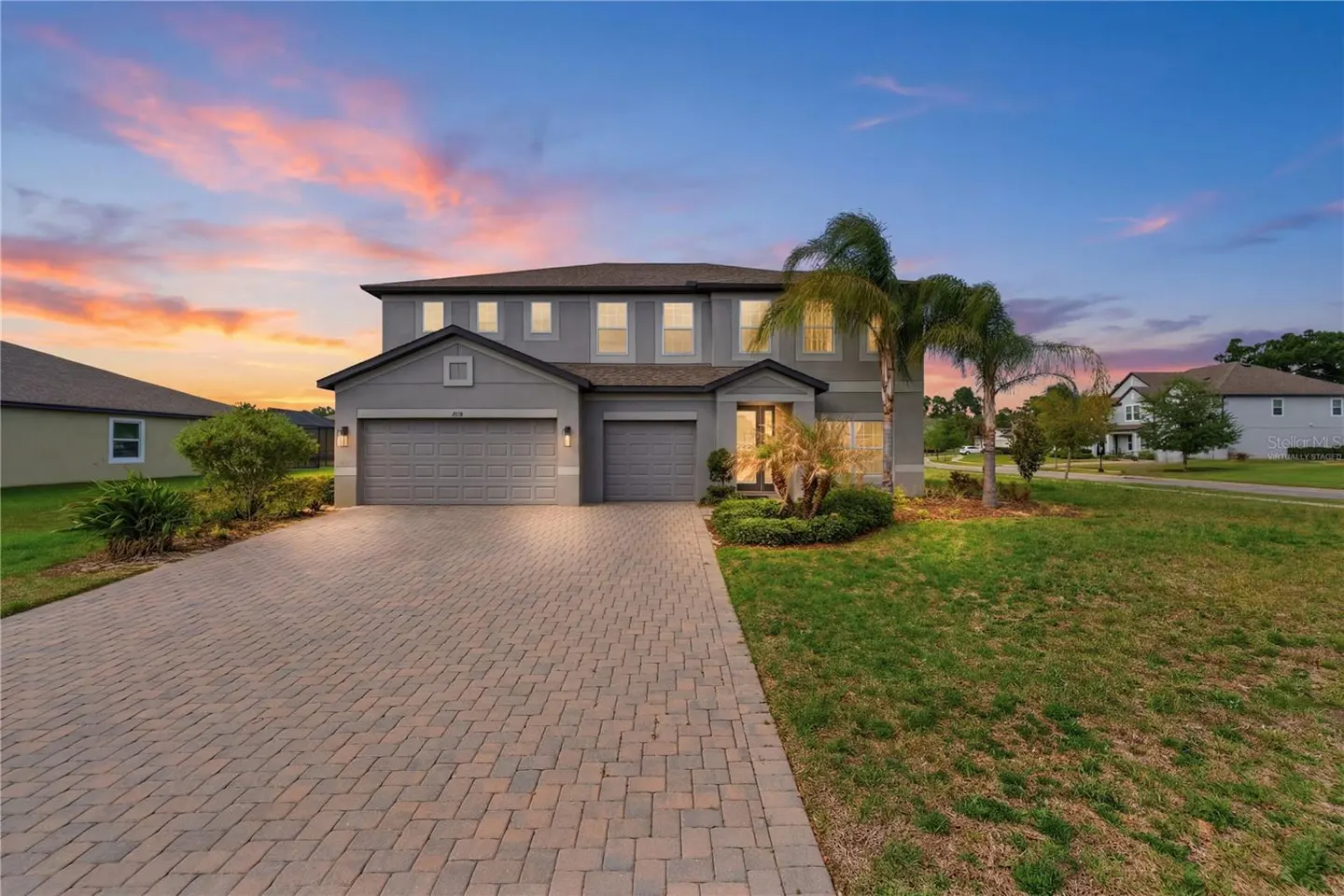 Two-story gray house with a brick driveway, green lawn, and palm trees at sunset.