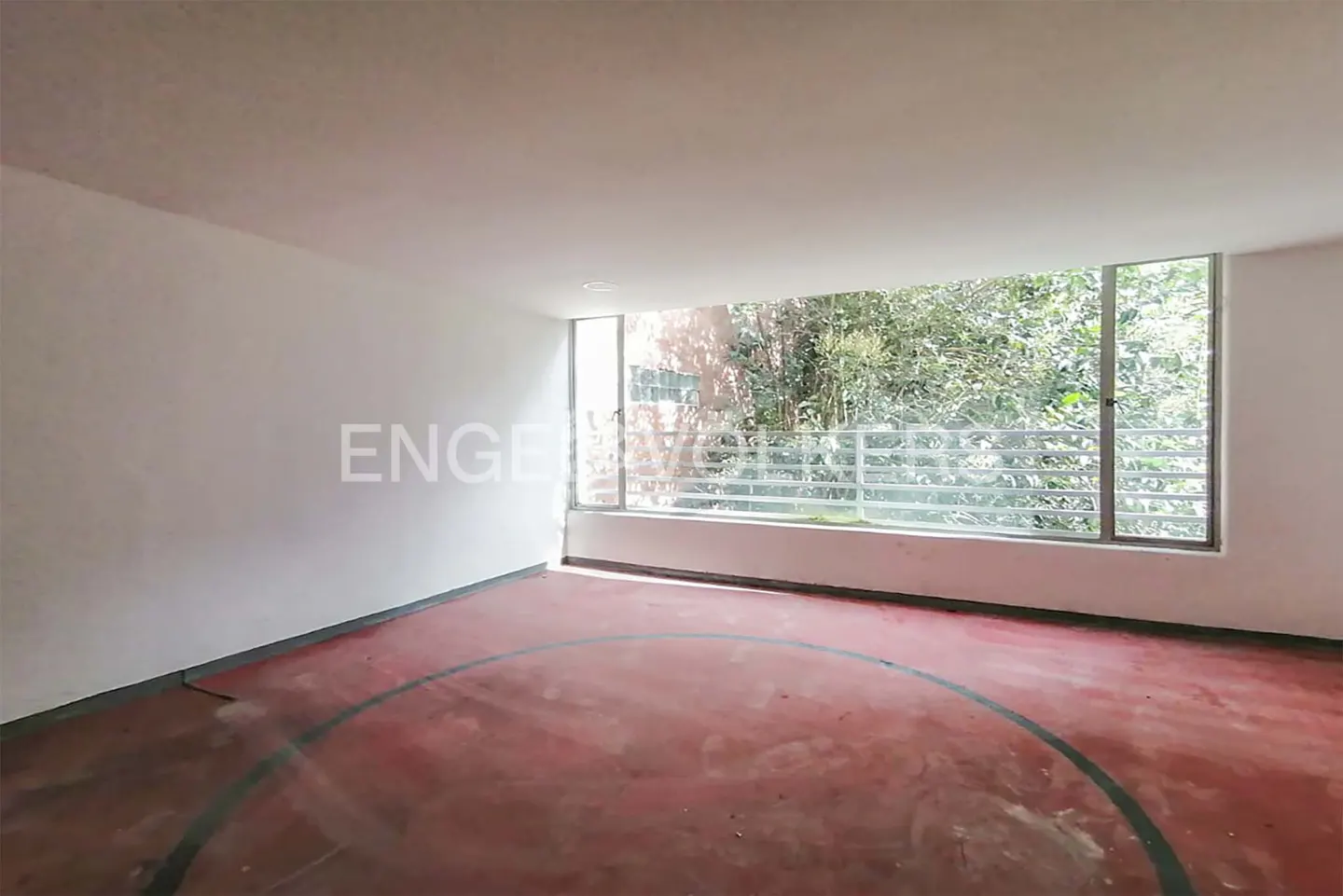 Empty room with white walls and a red floor with a black circle. A large window shows green trees outside.