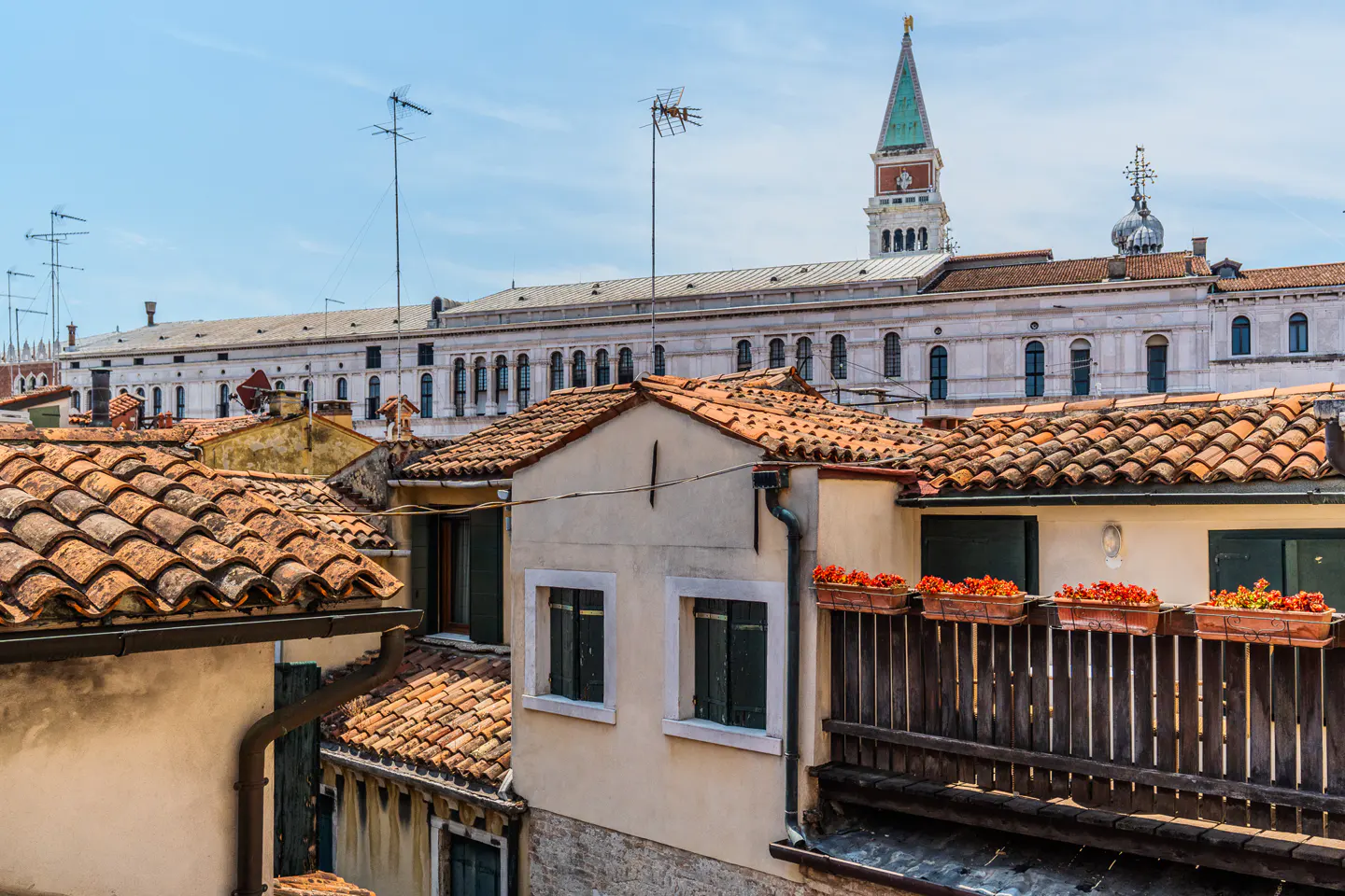 Rooftop view in Venice with terracotta tiles, a wooden balcony with red flowers, and a historic bell tower in the distance.