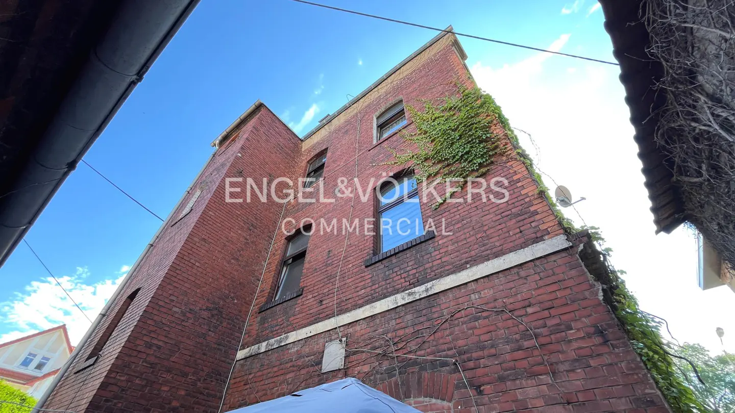 Low-angle shot of a red brick commercial building with green vines, under a blue sky. The Engel & Volkers logo is superimposed on the building.