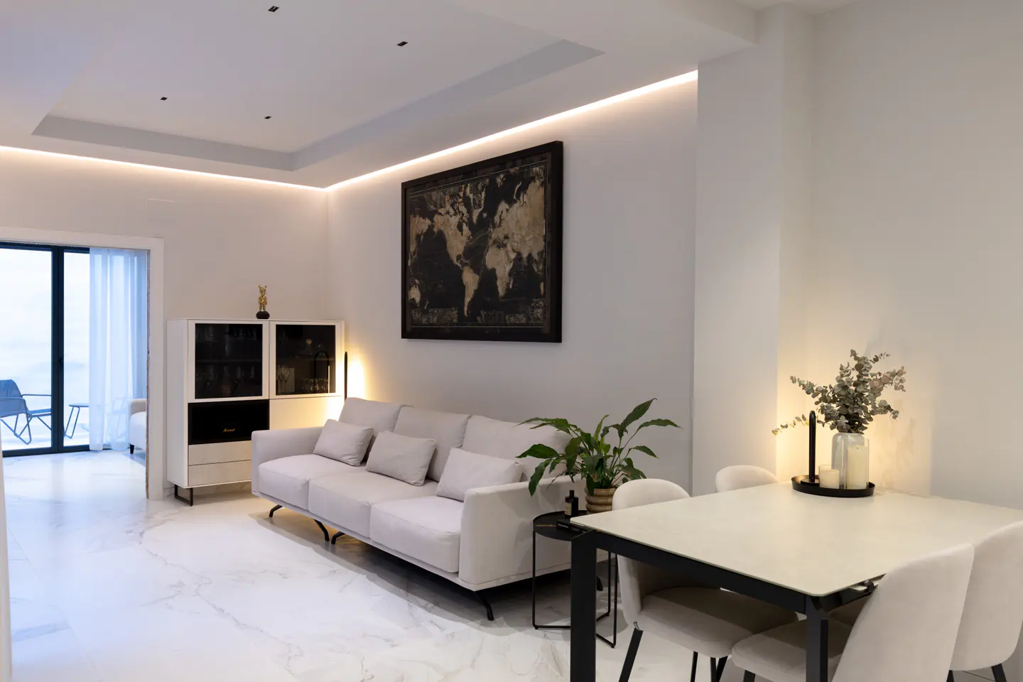 Bright, modern living room with white walls, marble floors, and recessed lighting. A light sofa sits beneath a world map. Dining table with four chairs.