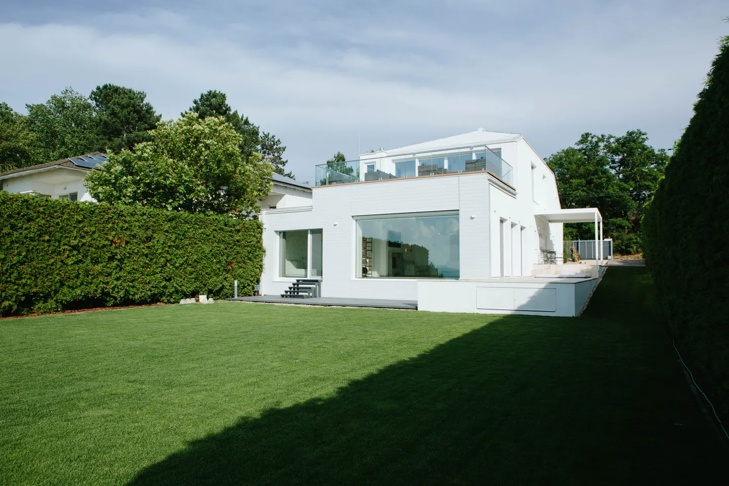 Modern white house with a flat roof and glass balcony, surrounded by green lawn and trees.