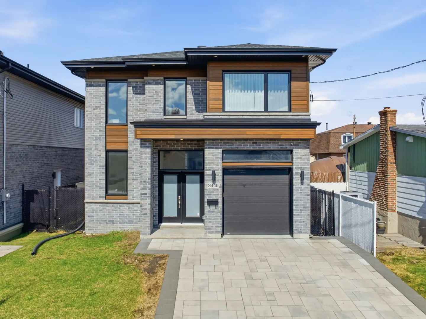 Two-story modern home with gray brick, wood accents, black trim, and a gray garage door. A paved driveway leads to the house.