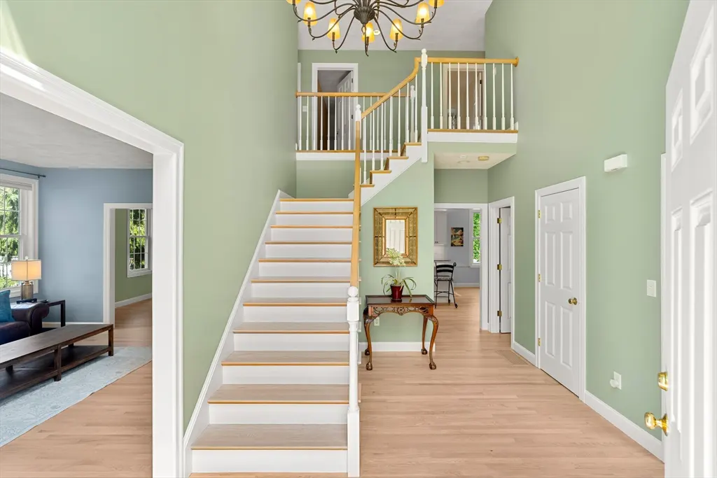 Light green foyer with white stairs, a wooden banister, and a chandelier. A living room is visible through an open doorway.