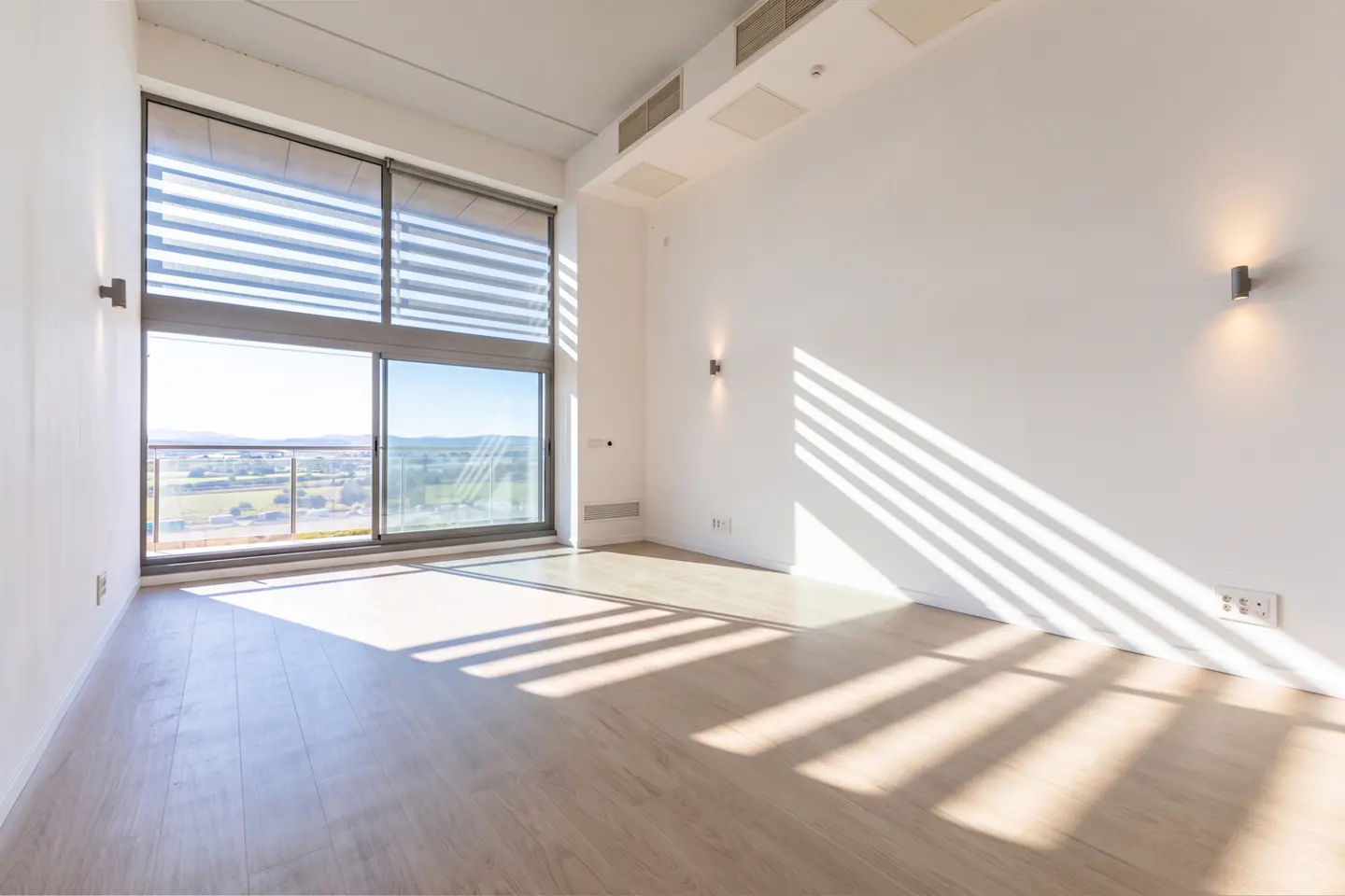 Bright, empty room with white walls, light wood floors, and a large window with blinds. Sunlight streams in, creating shadows on the floor.