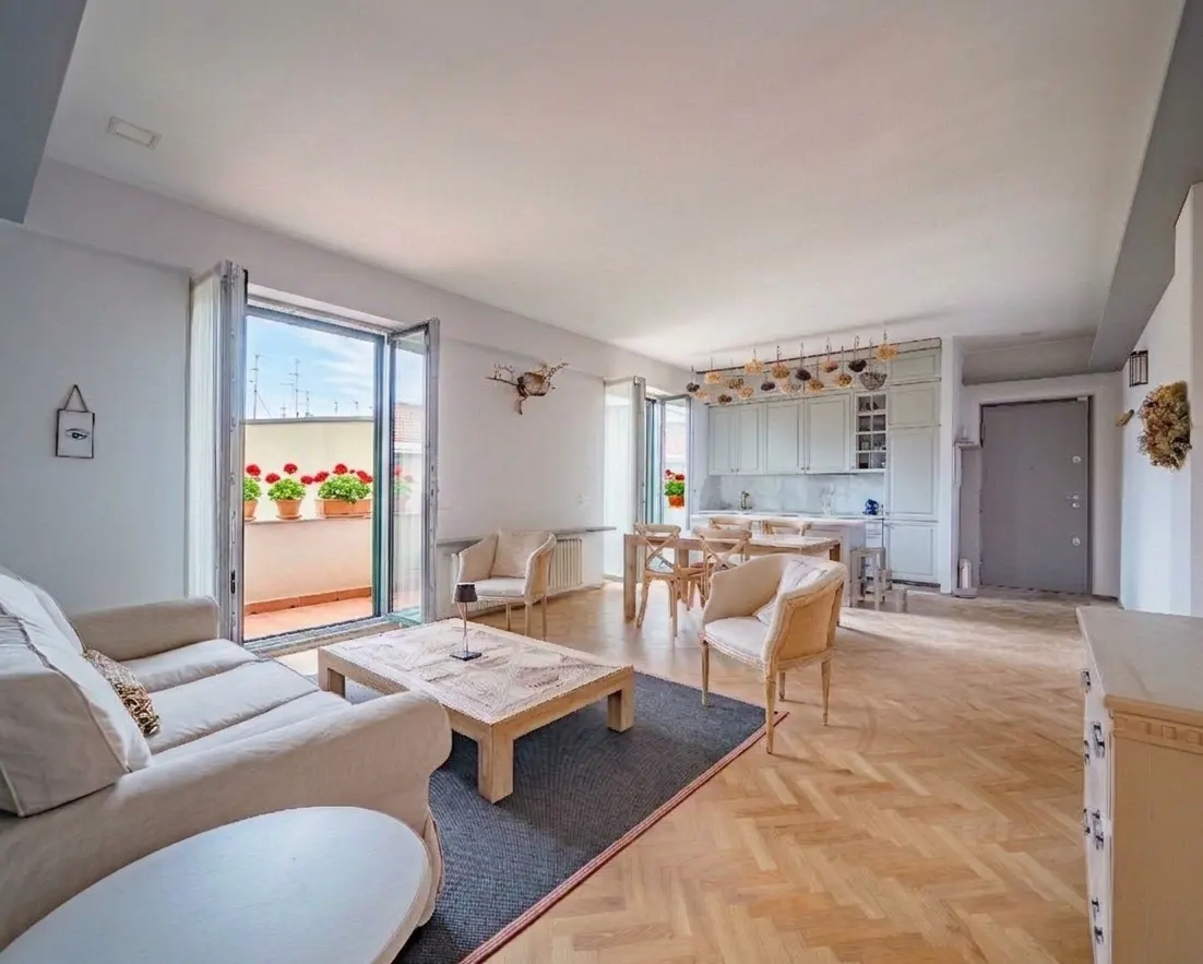 Bright, open-concept living space with a cream sofa, chairs, and a wooden coffee table on a gray rug. Balcony with red flowers visible.