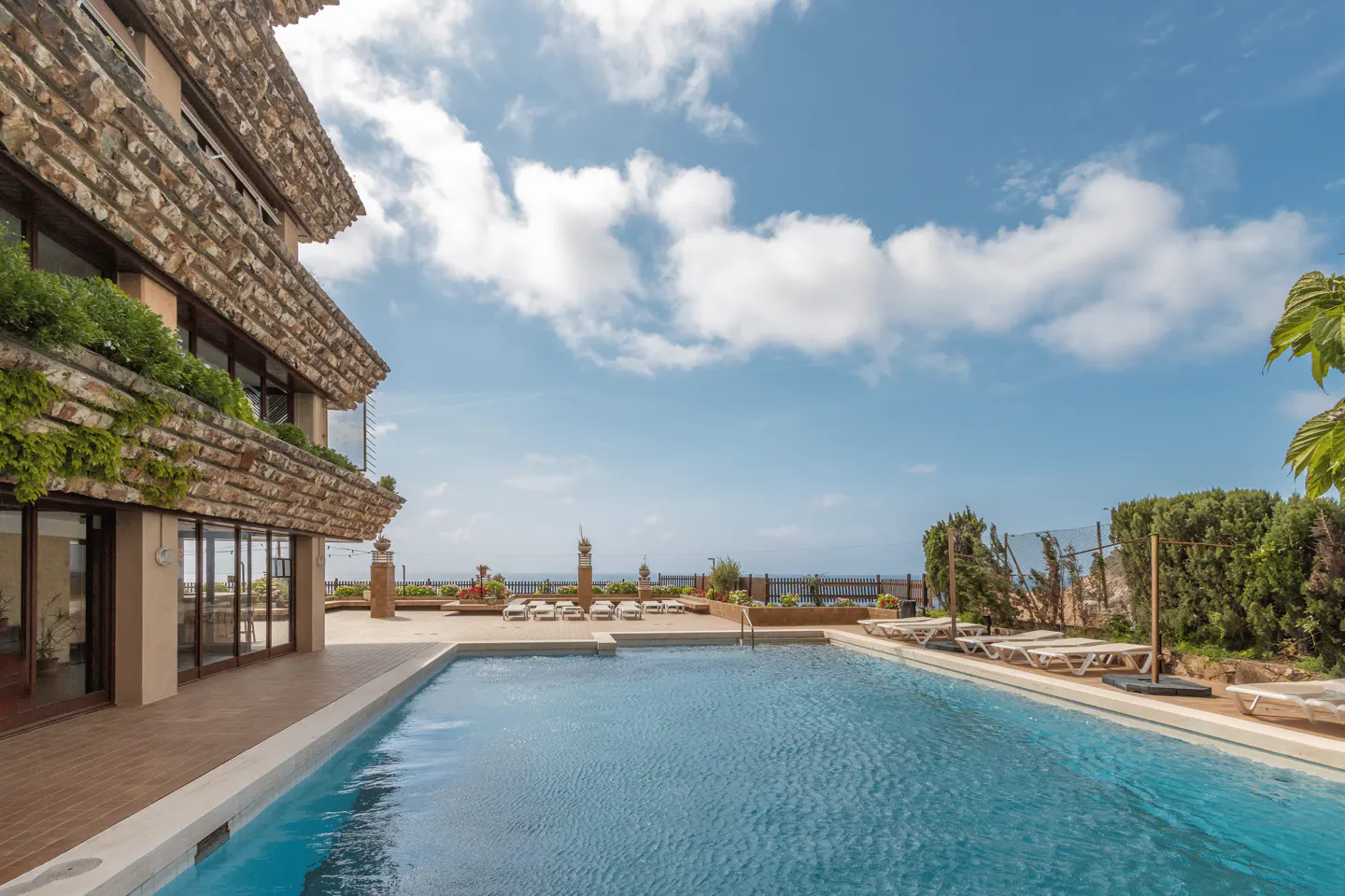 A swimming pool with blue water sits beside a multi-story stone building with greenery under a bright, cloudy blue sky.