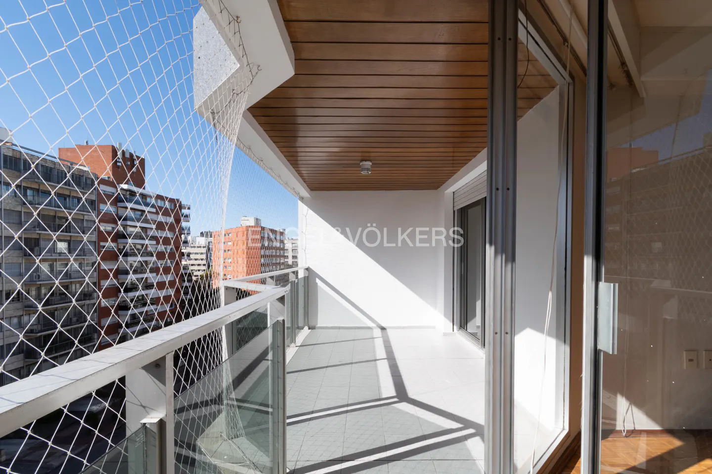 Balcony view with white railings, safety net, and city buildings in the background. The ceiling is wood and the floor is tiled.