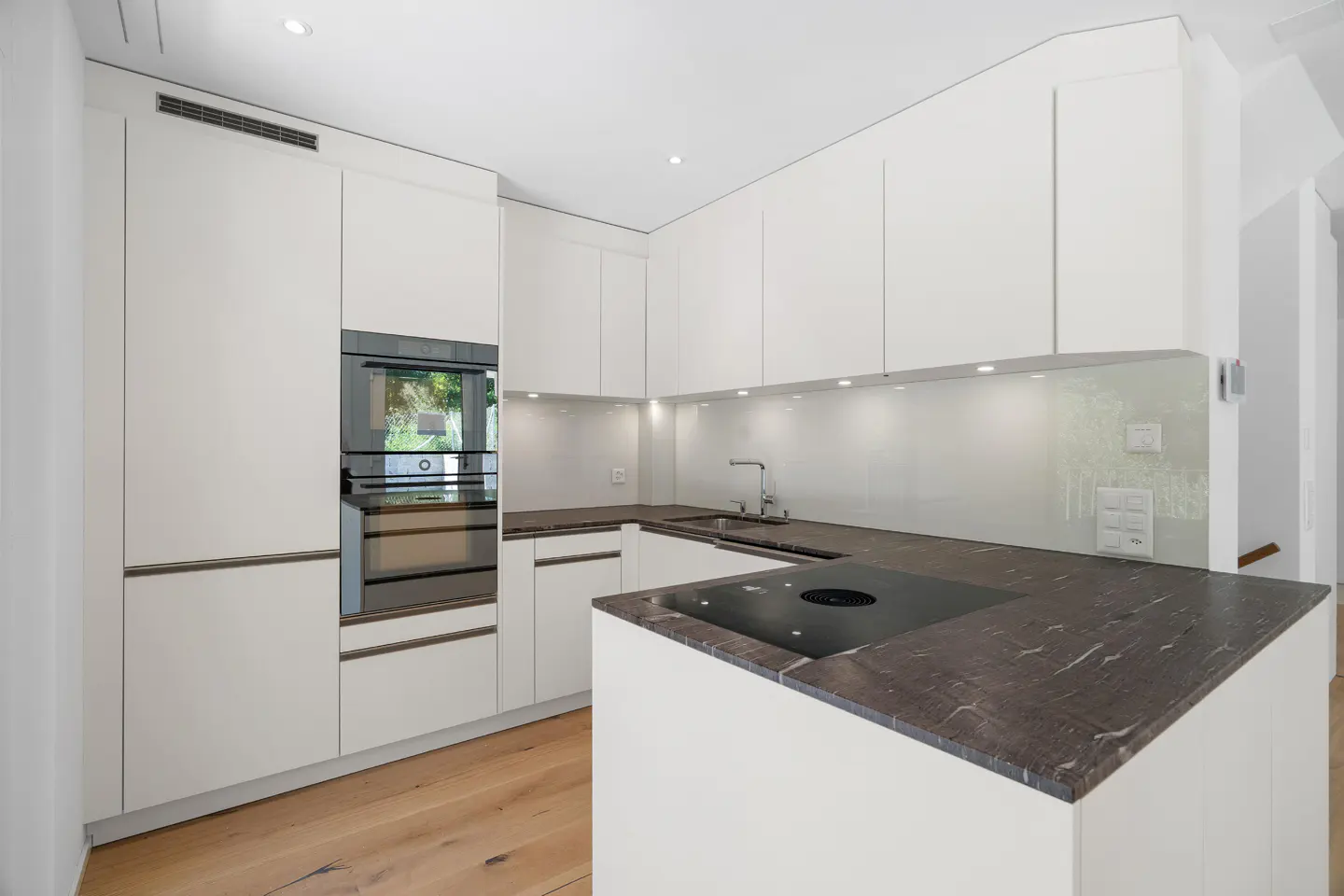 Modern white kitchen with dark marble countertops, built-in oven, and light wood flooring.