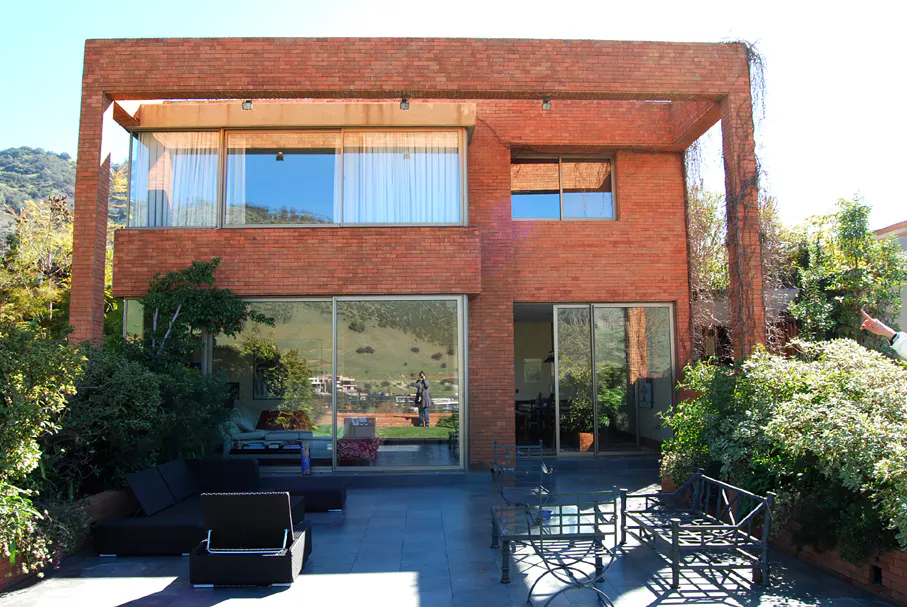 Two-story brick house with large windows and a patio with outdoor furniture. Greenery surrounds the house.