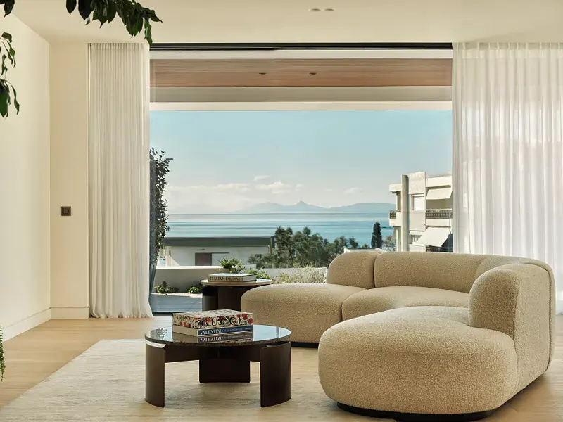 Bright living room with a beige, curved sofa, round tables, and a large window overlooking the ocean. White curtains frame the view.
