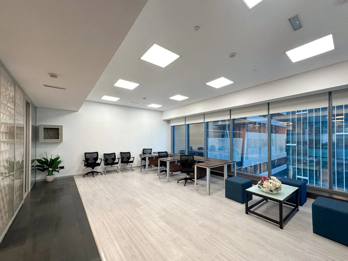 Bright office space with large windows, desks, chairs, and blue ottomans. A plant sits near a textured glass wall.