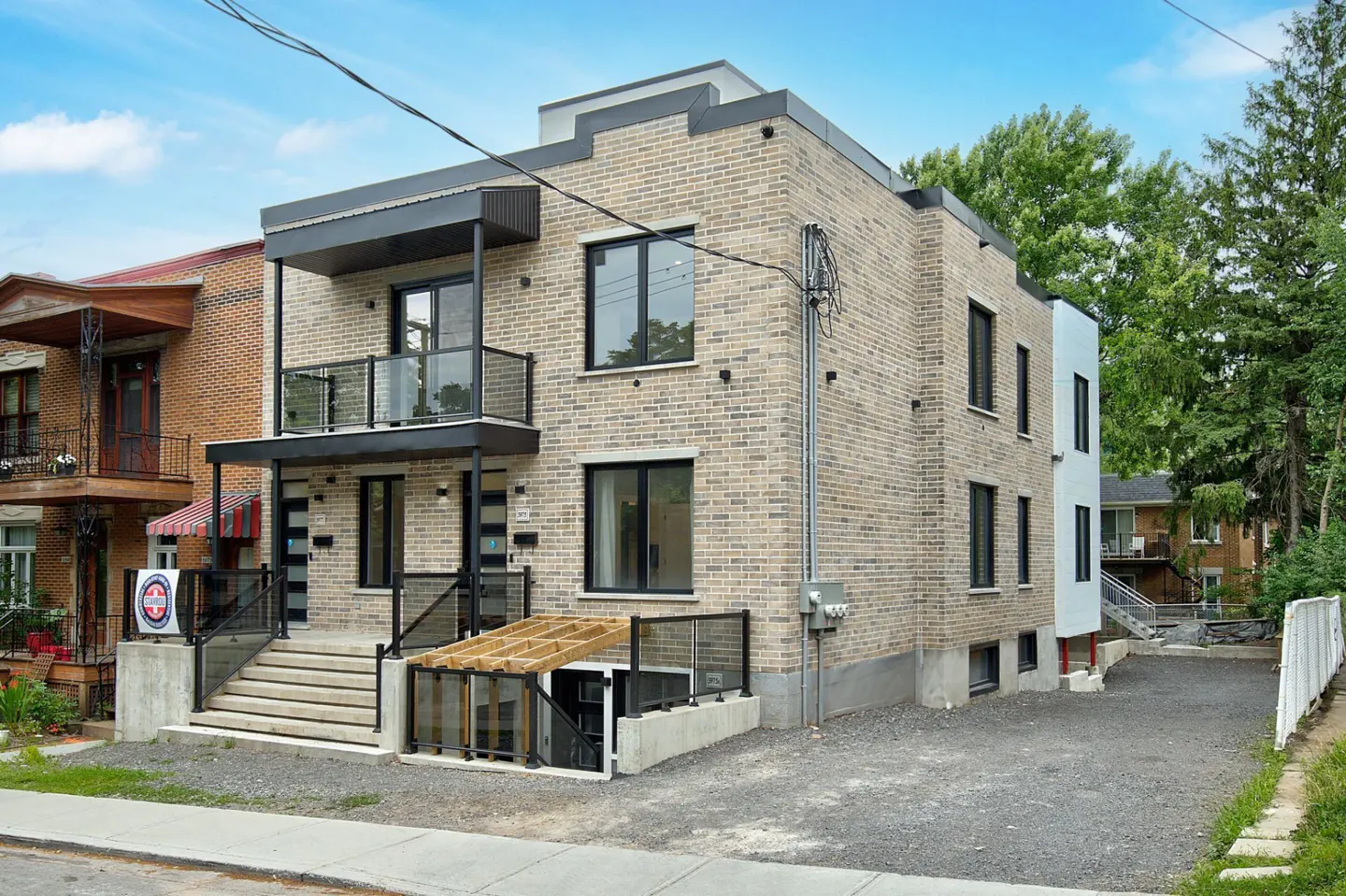 Two-story tan brick building with black trim, balconies, and a stairway leading to the entrance. A basement entrance is visible on the side.