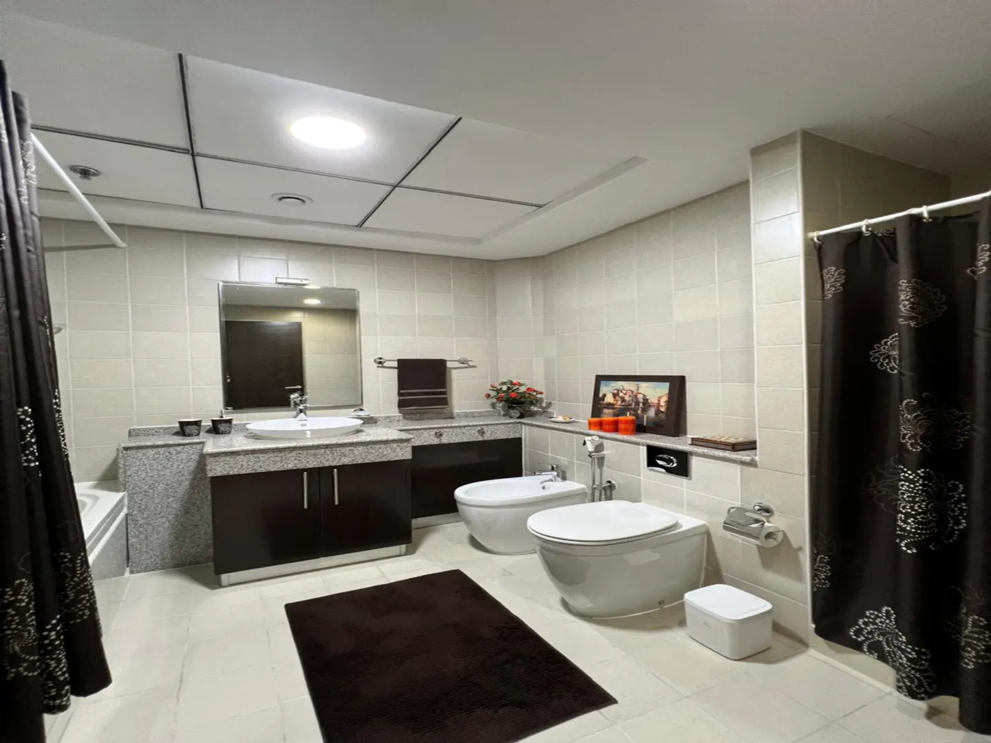 A bathroom with a sink, toilet, bidet, and a dark brown rug. The walls are tiled and there is a dark brown shower curtain.