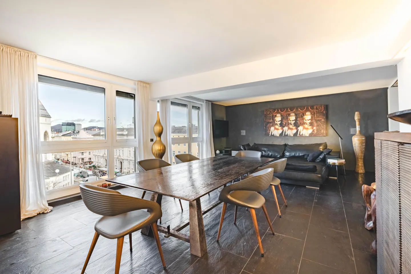 Modern dining room with a long, dark wood table and six chairs. Large windows offer a city view. A black leather sofa sits against a gray wall with art.