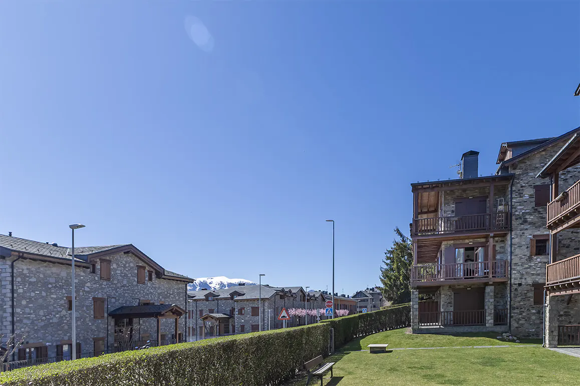 Stone buildings with wooden balconies under a clear blue sky. A green hedge and lawn are in the foreground. Snow-capped mountains are visible in the distance.