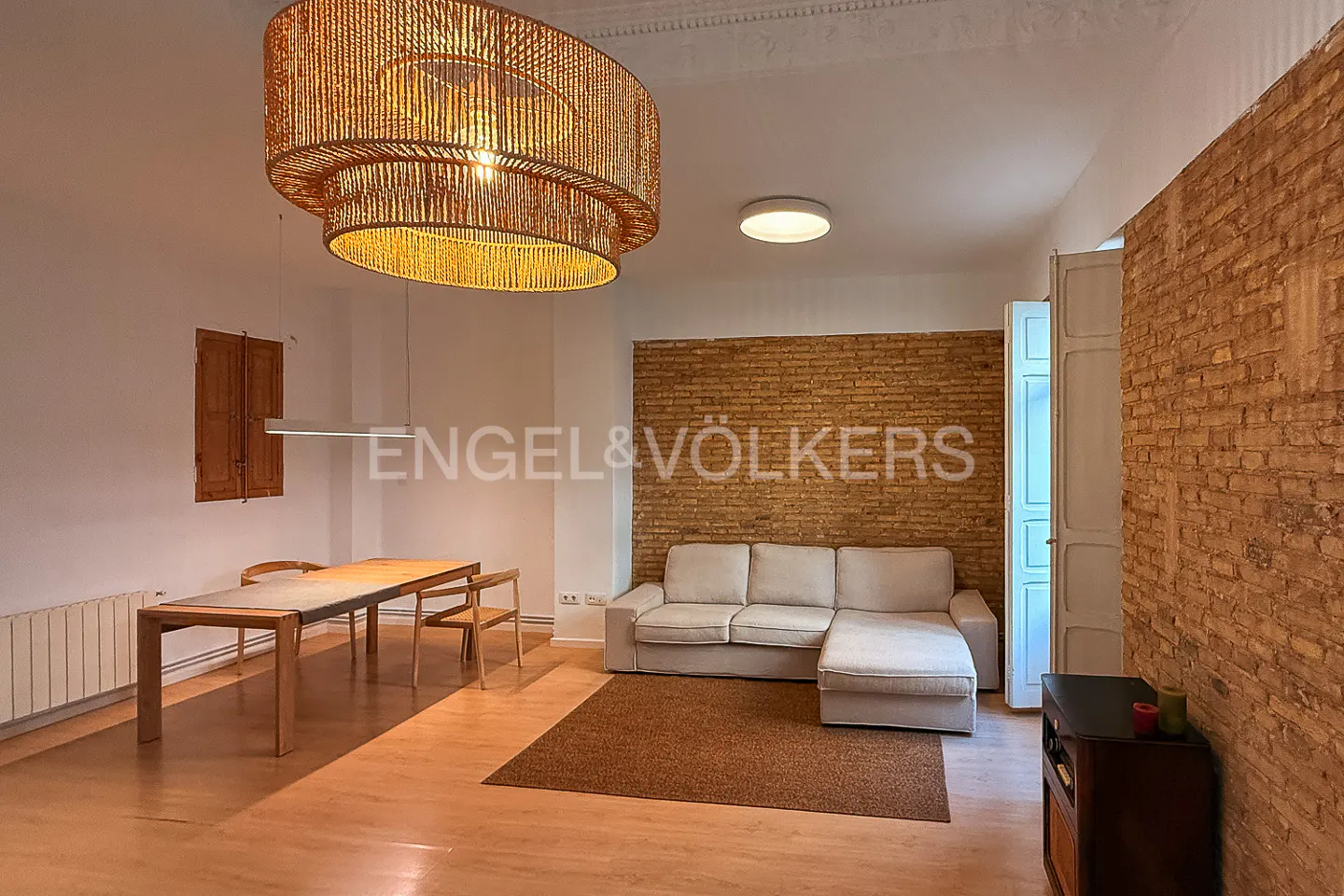 Living room with a light sofa, brick wall, wooden table, and a large, round, woven chandelier.