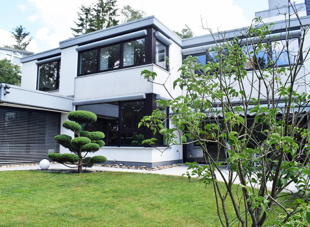 Modern white house with black framed windows, green lawn, and sculpted tree.