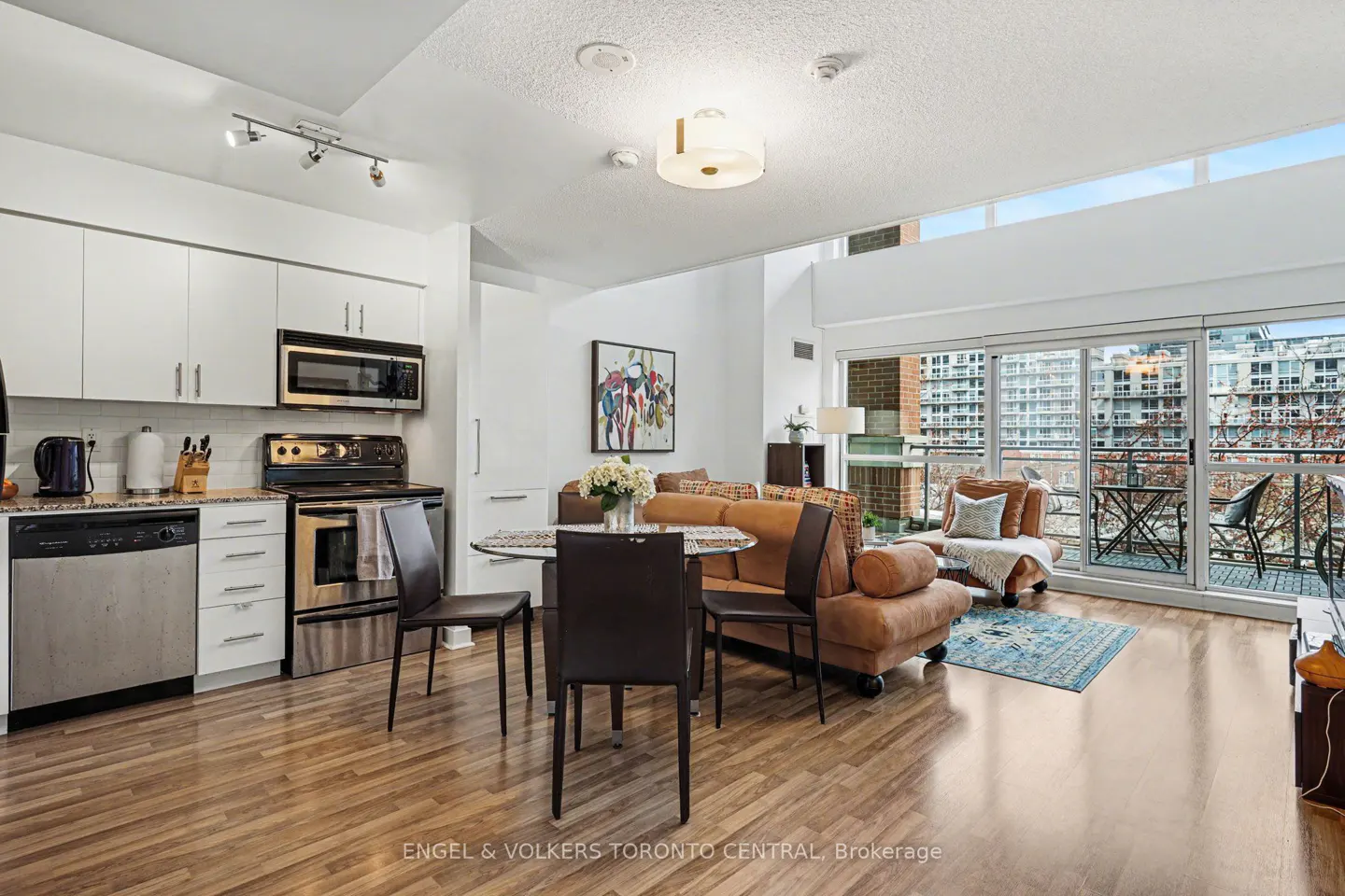 Bright condo interior with white kitchen cabinets, stainless steel appliances, and a brown leather sofa near a balcony.