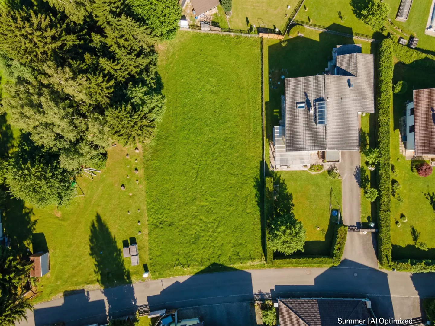 Aerial view of a large, green, grassy lot next to a house with a gray roof and a driveway. Trees surround the property.
