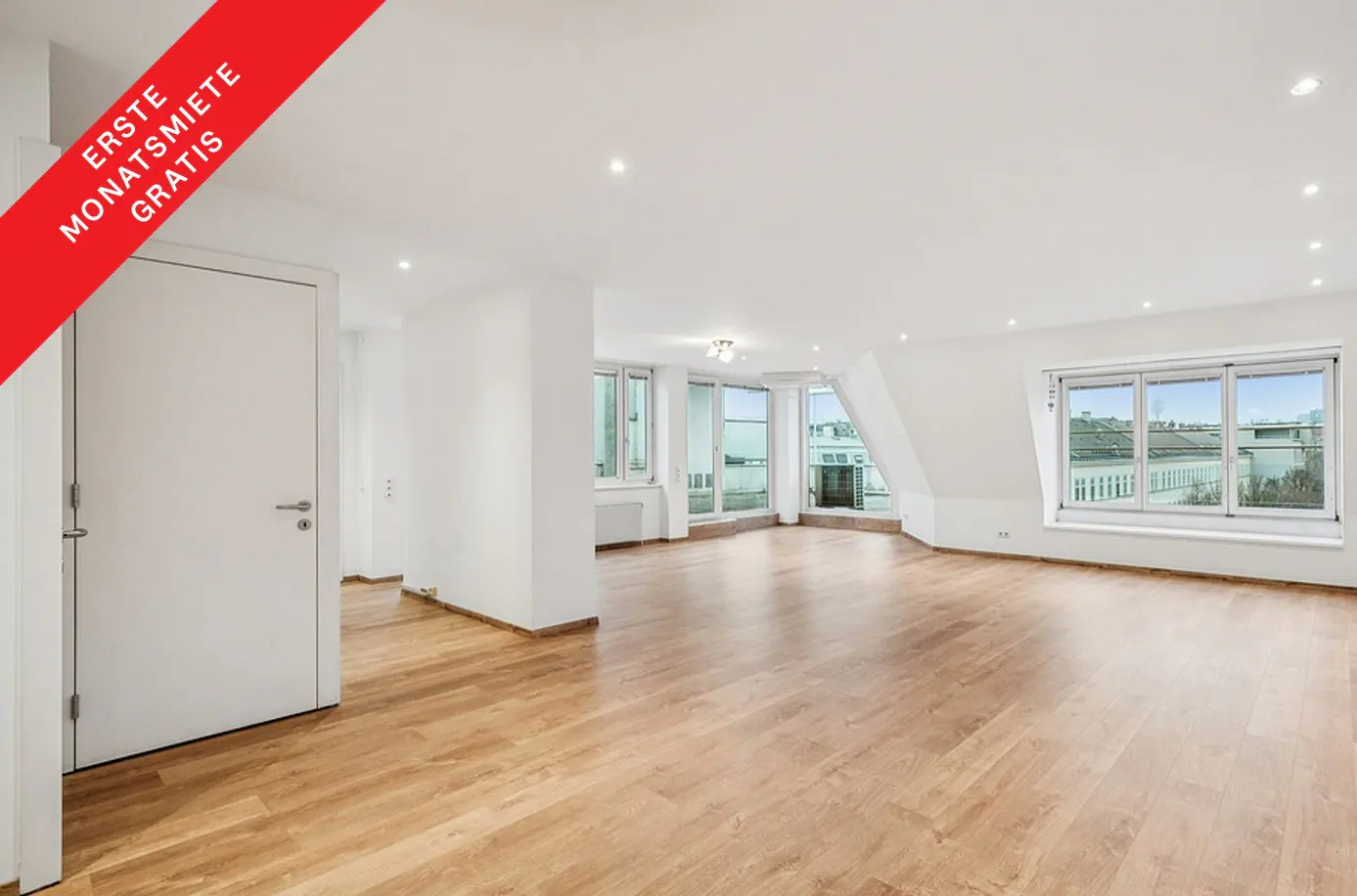Bright, empty room with hardwood floors, white walls, and large windows overlooking a city. A red banner in the corner reads "Erste Monatsmiete Gratis."