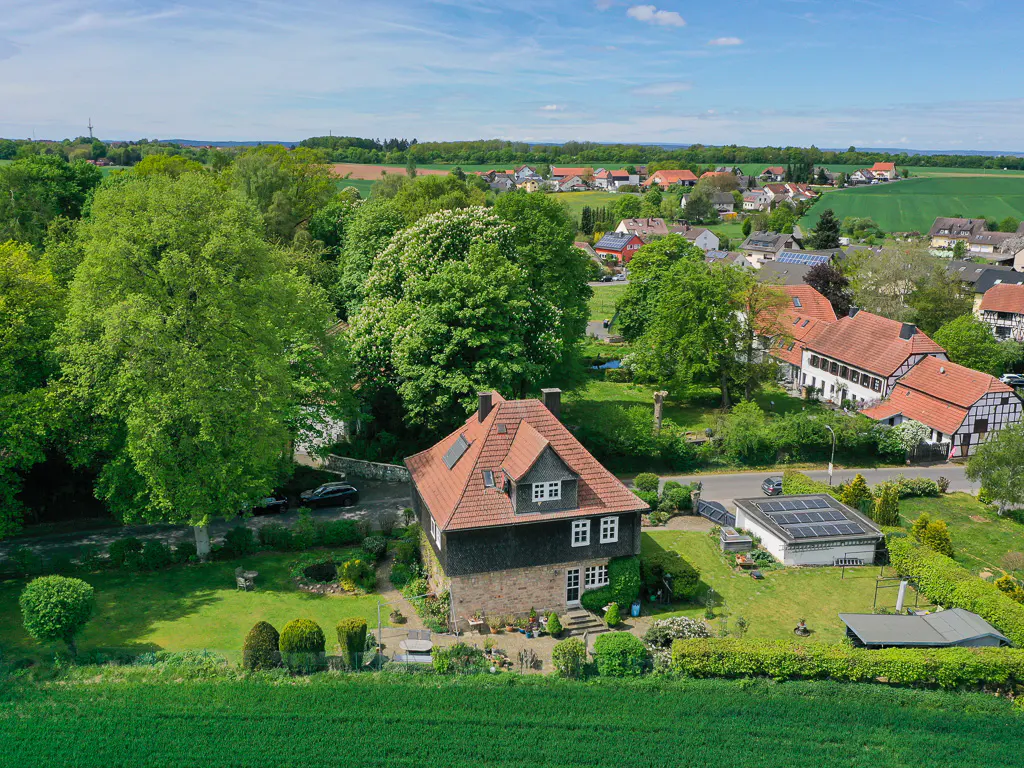Aerial view of a two-story house with a red tile roof and stone base, surrounded by lush trees and a green lawn.