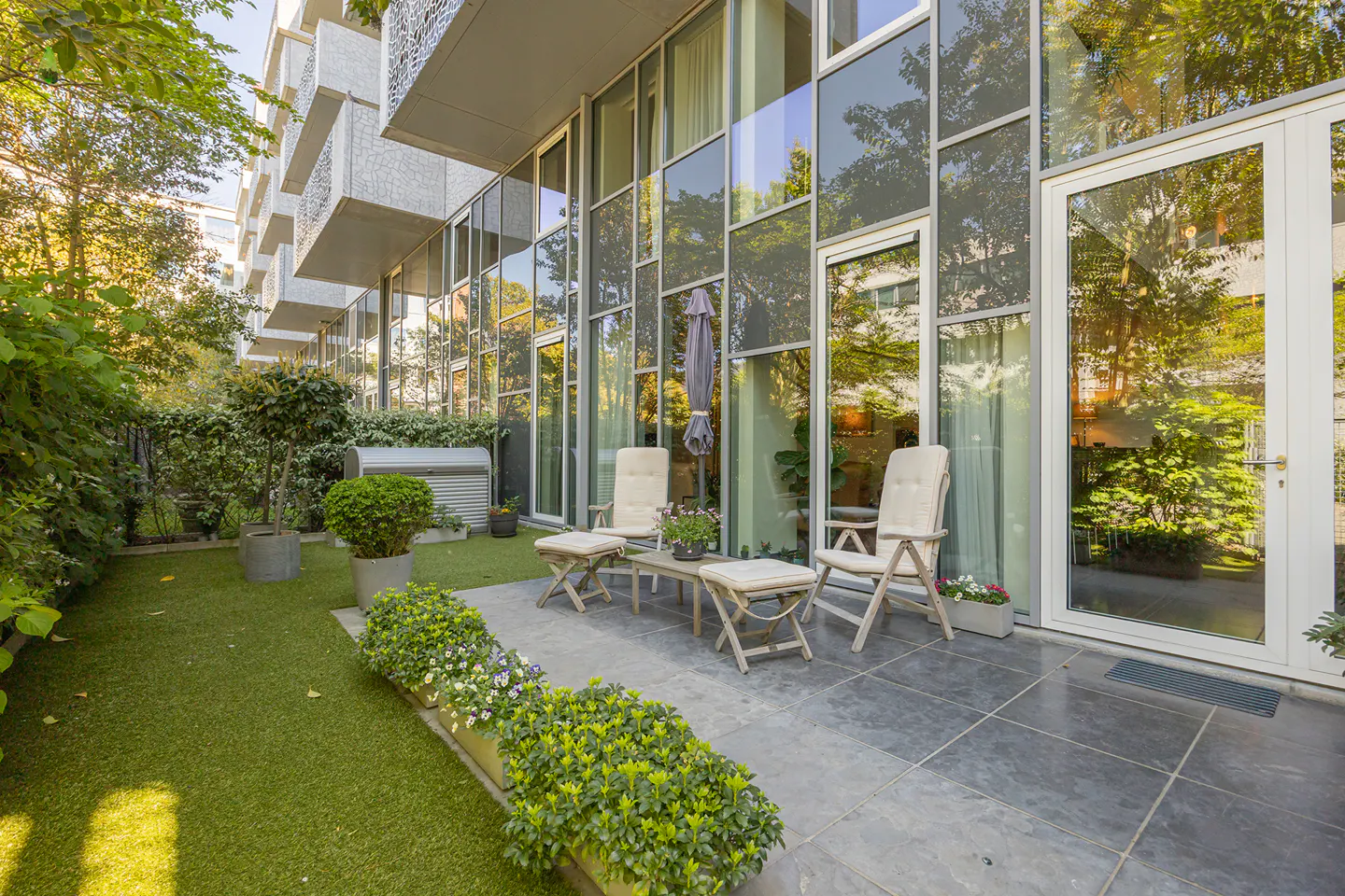 Outdoor patio with chairs, table, and green space. Modern building with large glass windows reflecting trees.