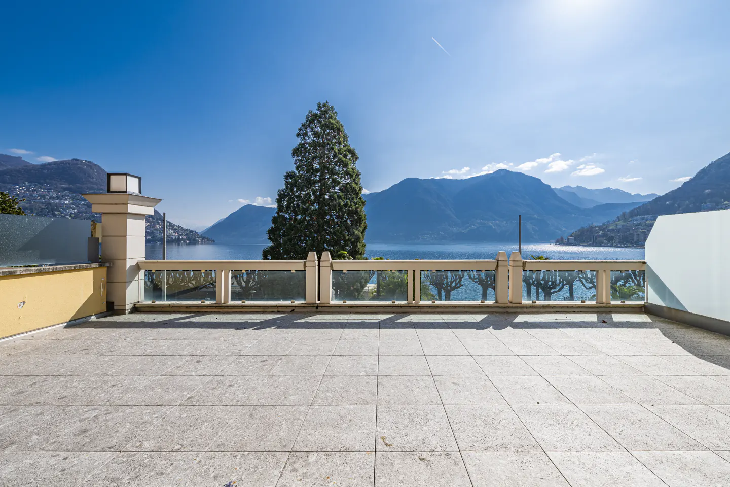 Balcony view of a lake and mountains. The balcony has a stone floor and glass railing. A tall tree is in the background.