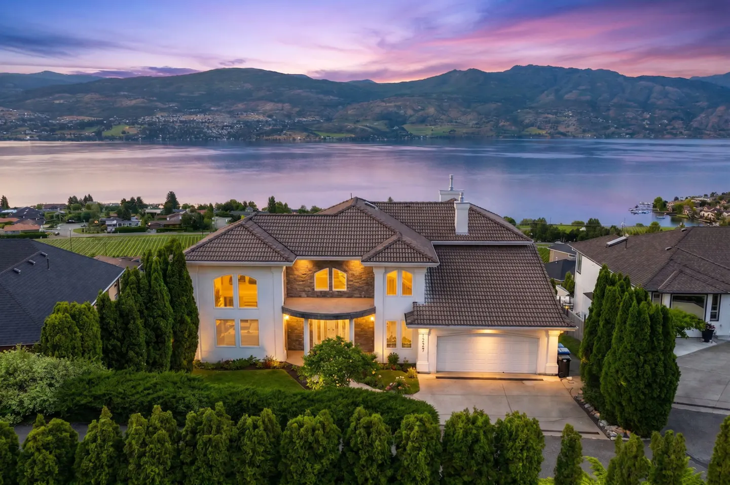 Luxury two-story home with a brown tile roof, illuminated windows, and a driveway, overlooking a lake and mountains at dusk.