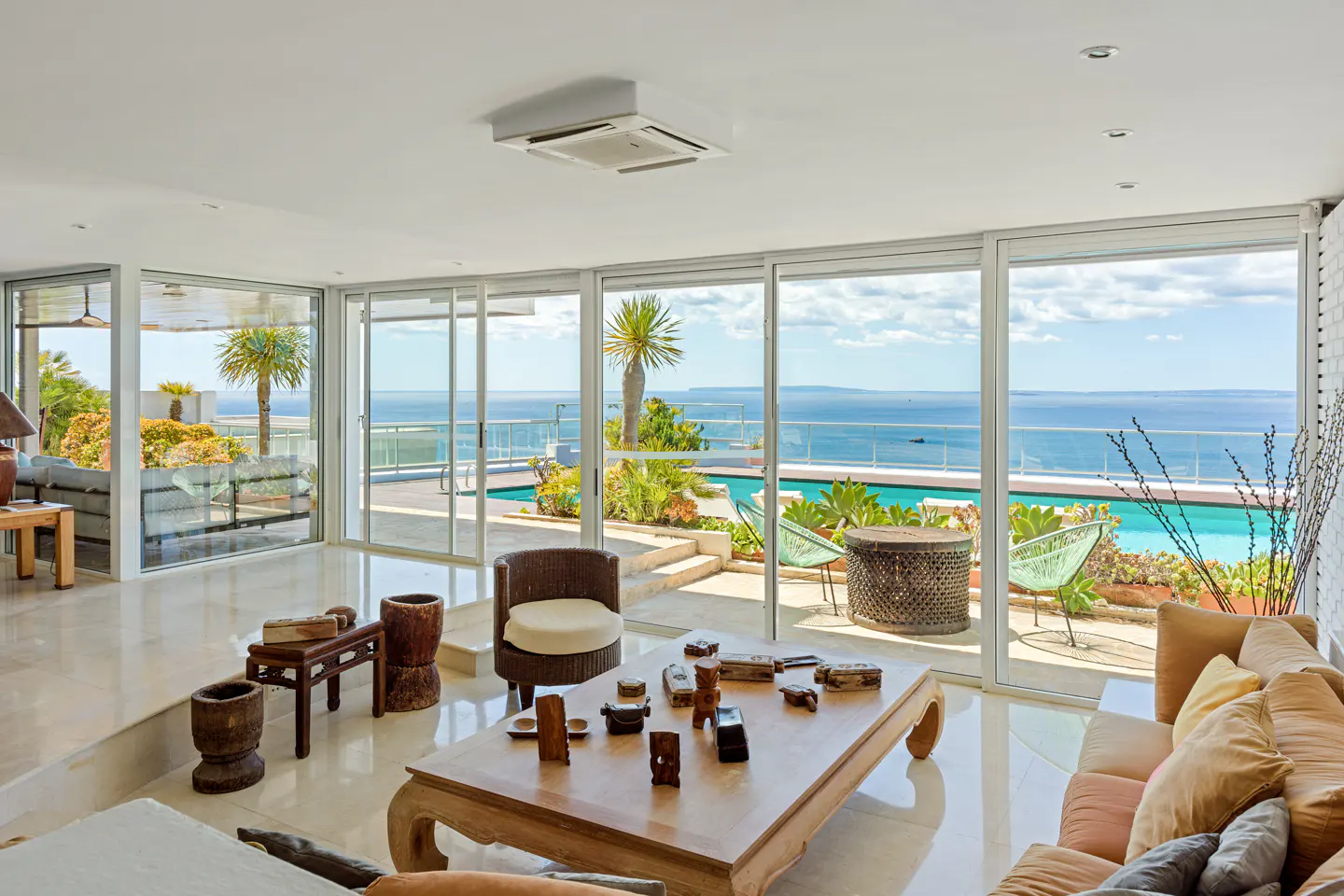 Bright living room with a large window view of a pool and ocean. A wooden table and wicker chair sit in the foreground.