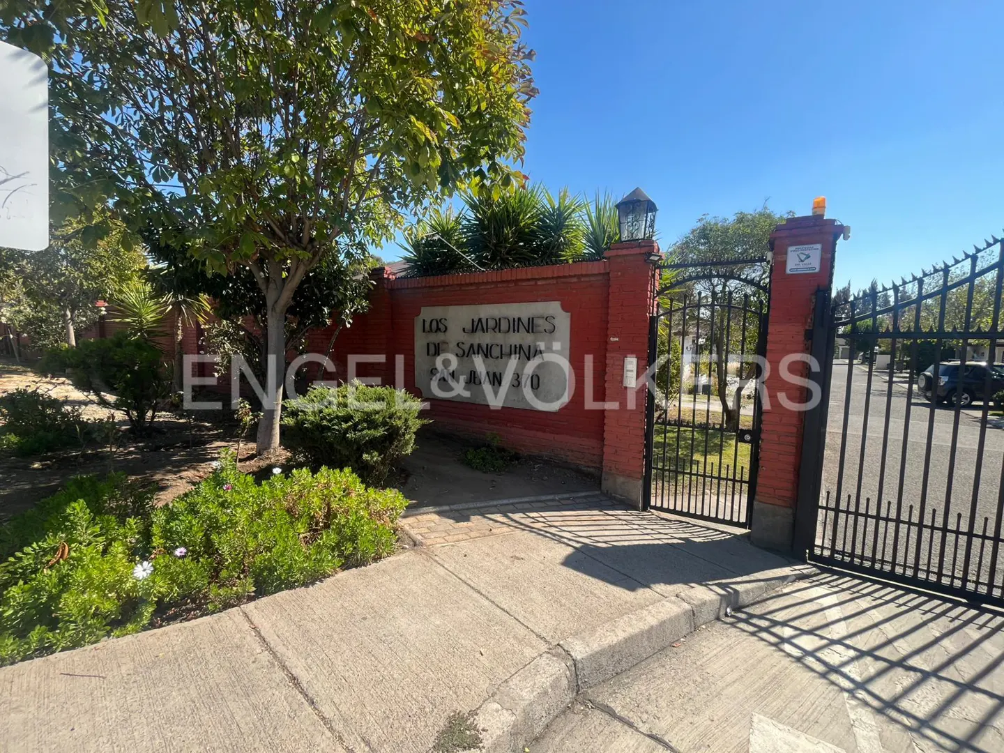 Exterior view of "Los Jardines de Sanchina" entrance with Engel & Völkers sign on a red brick wall and black iron gate.