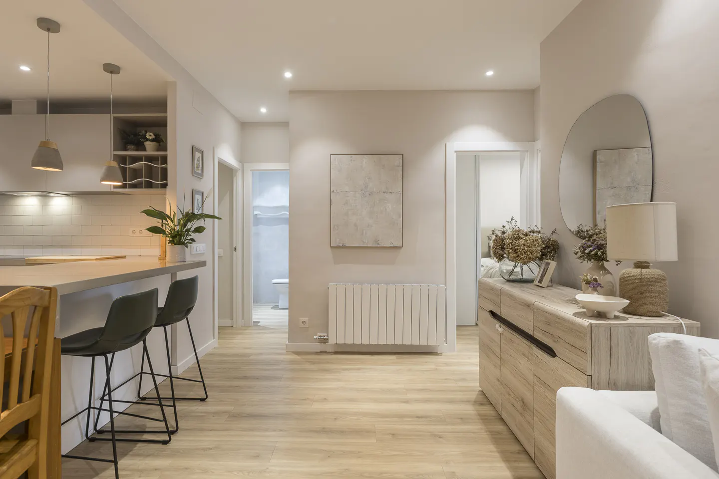 A bright, modern apartment interior with a kitchen island, bar stools, and a hallway leading to other rooms. Neutral color palette.