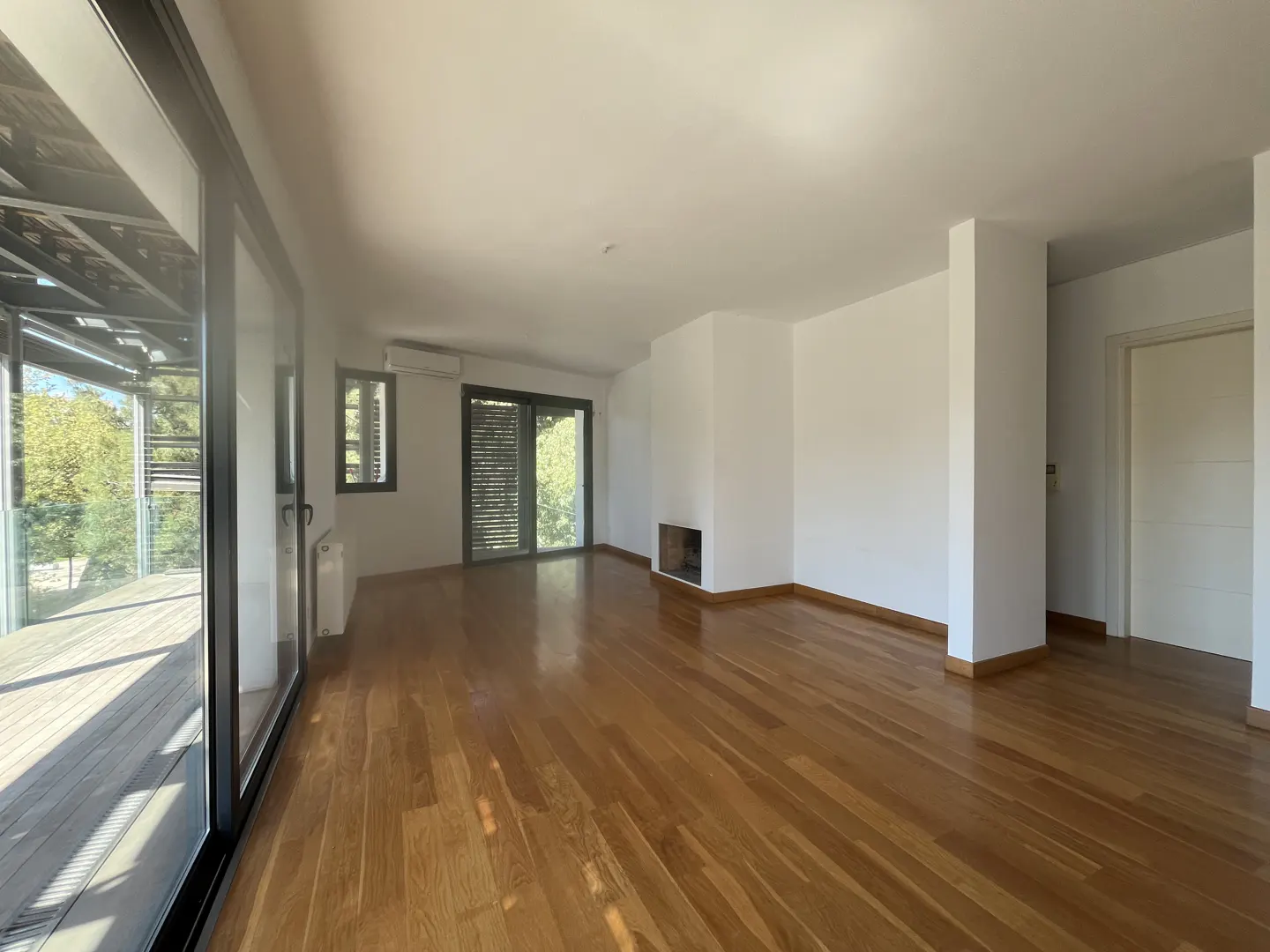 Bright, empty living room with hardwood floors, white walls, and a fireplace. Large windows and sliding doors lead to an outdoor deck.