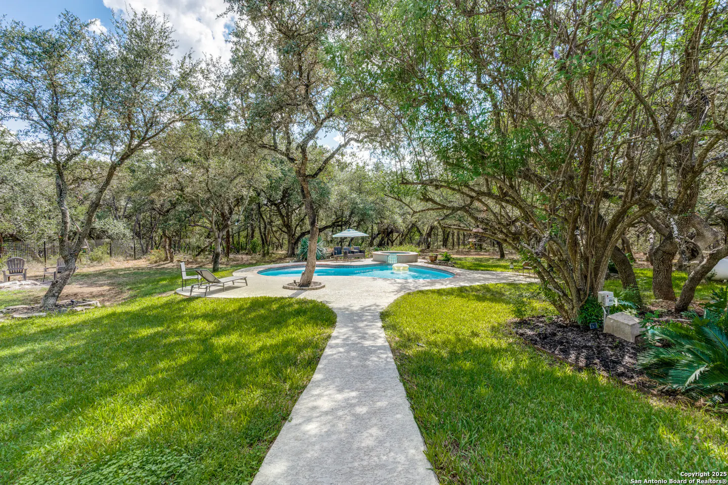 A backyard with a pool, green grass, and trees. A concrete path leads to the pool area with lounge chairs and an umbrella.
