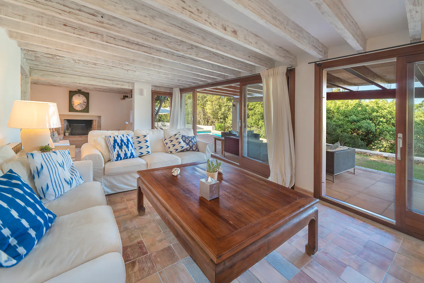 Living room with white sofas, blue pillows, and a large wooden coffee table. Sliding glass doors lead to a patio with green trees in the background.