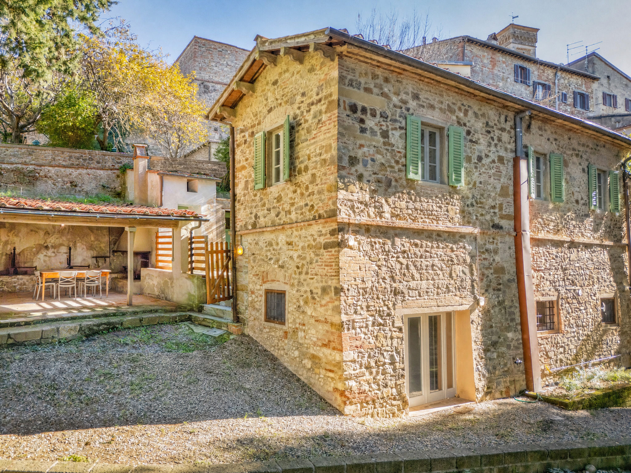 A rustic stone house in Italy with green shutters, a gravel yard, and a covered patio with outdoor dining furniture.