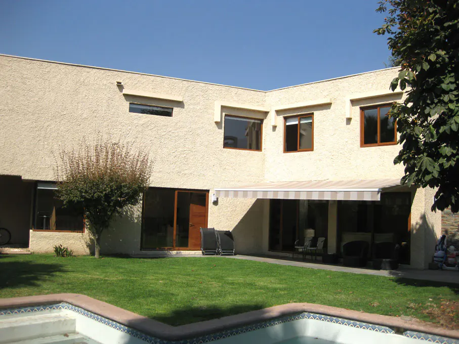 Backyard view of a two-story beige house with brown window frames, green lawn, and a pool in the foreground.