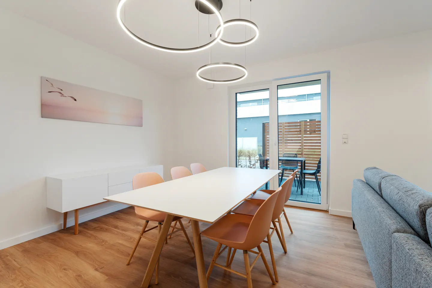 Bright dining room with a white table, peach chairs, and modern ring lights. A white cabinet and a painting of birds complete the scene.