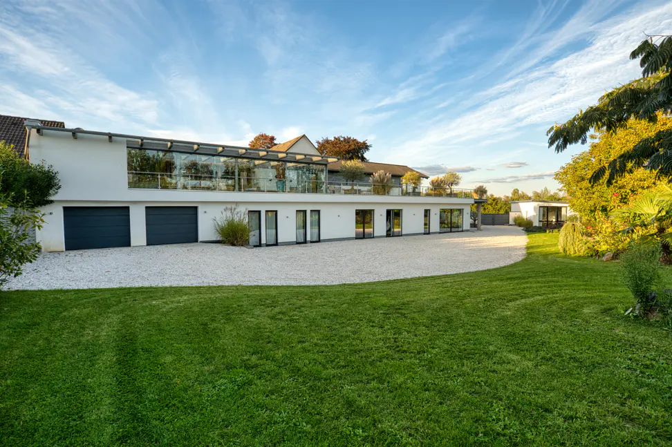 Modern white house with a glass-enclosed upper level, dark gray garage doors, and a gravel driveway leading to a green lawn.