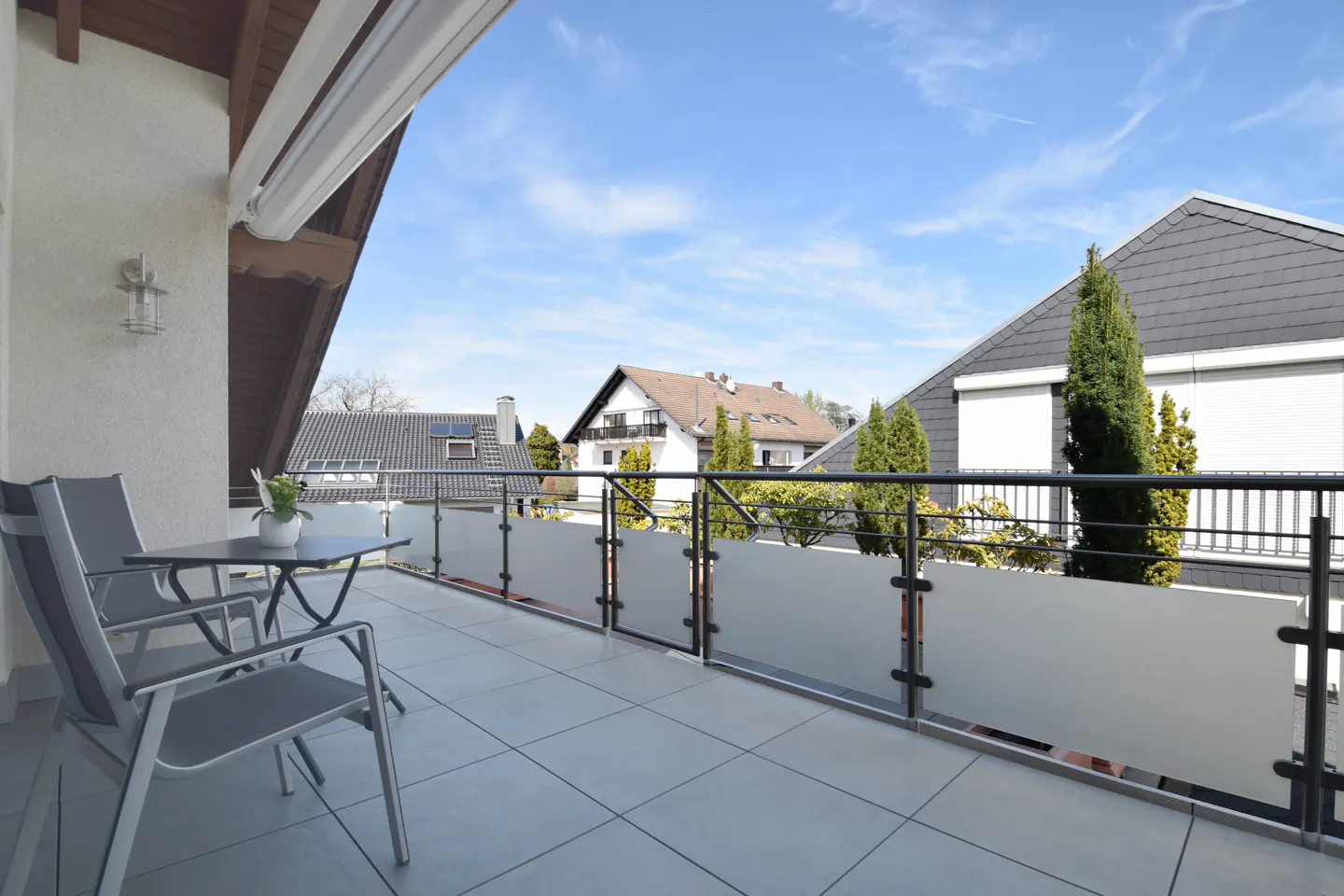 Balcony with gray tile floor, metal railing, table, and chairs. Houses and blue sky are visible in the background.