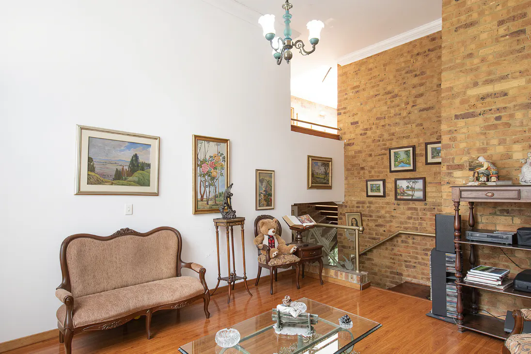Living room with a brown sofa, glass table, and brick wall. Paintings hang on the white and brick walls. A chandelier hangs from the ceiling.