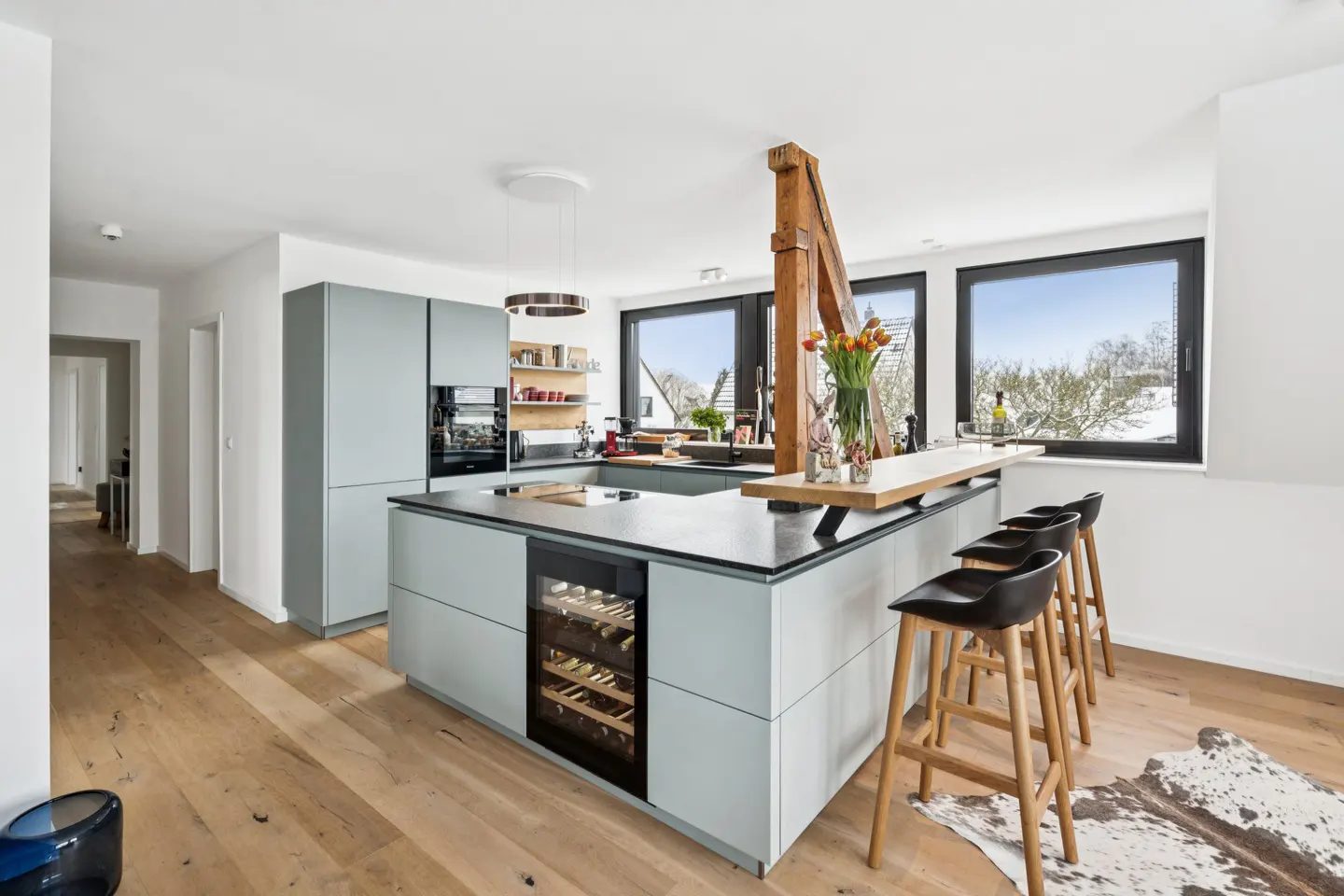 Bright kitchen with light blue cabinets, black countertops, and wood floors. Island with wine fridge and bar seating. Large windows offer natural light.