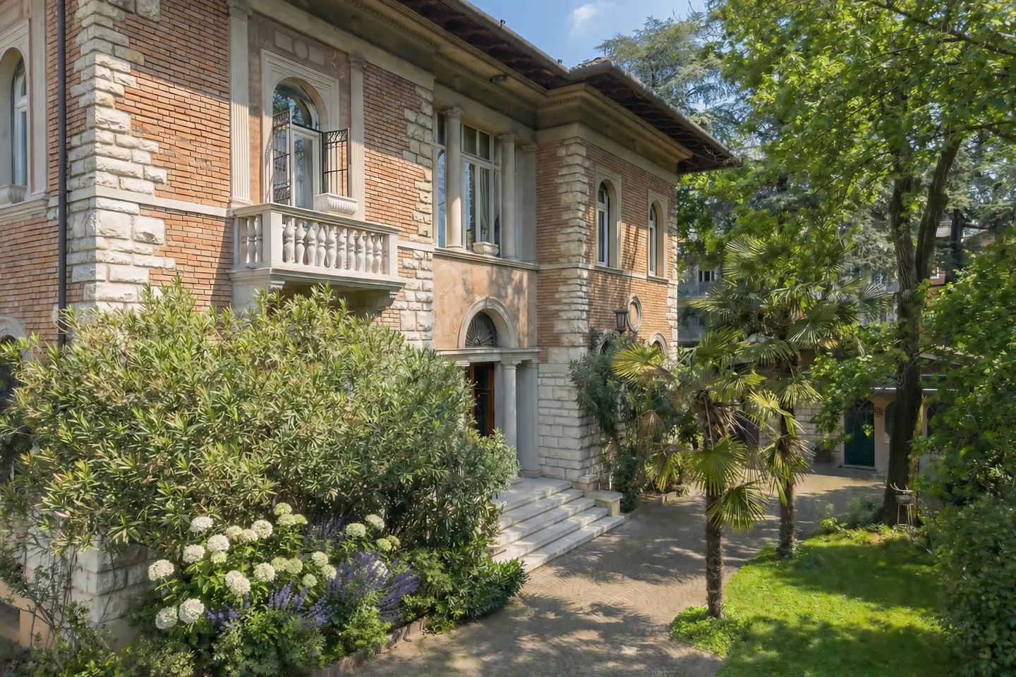 Exterior of a two-story brick and stone house with a balcony, steps, and lush green landscaping.