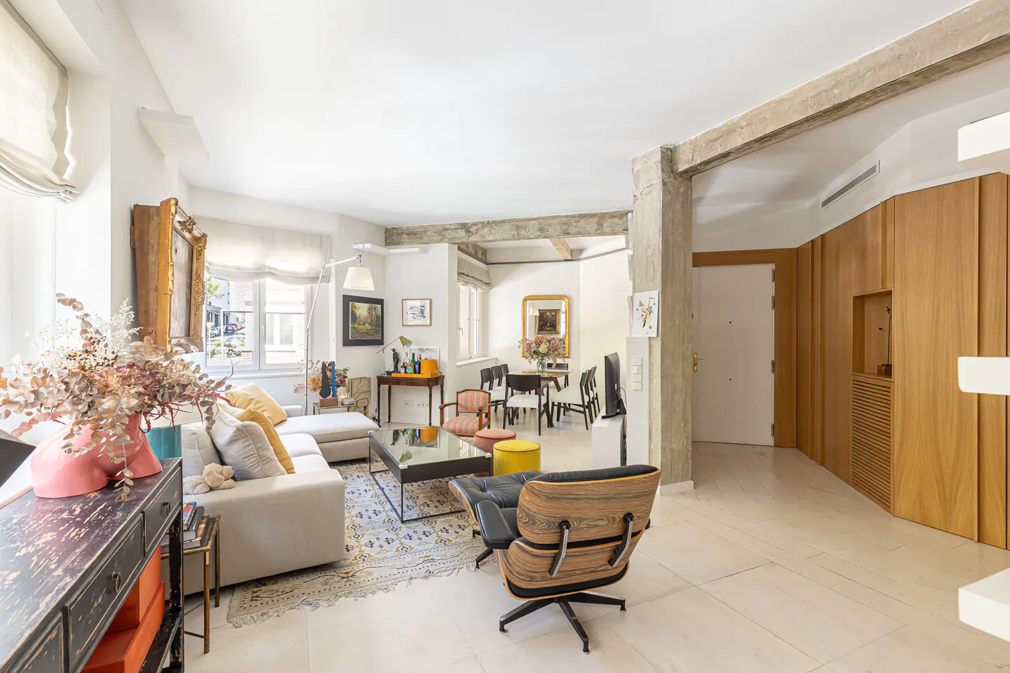 Bright, open living room with white walls, concrete beams, and wood accents. A white sofa, black coffee table, and Eames chair furnish the space.