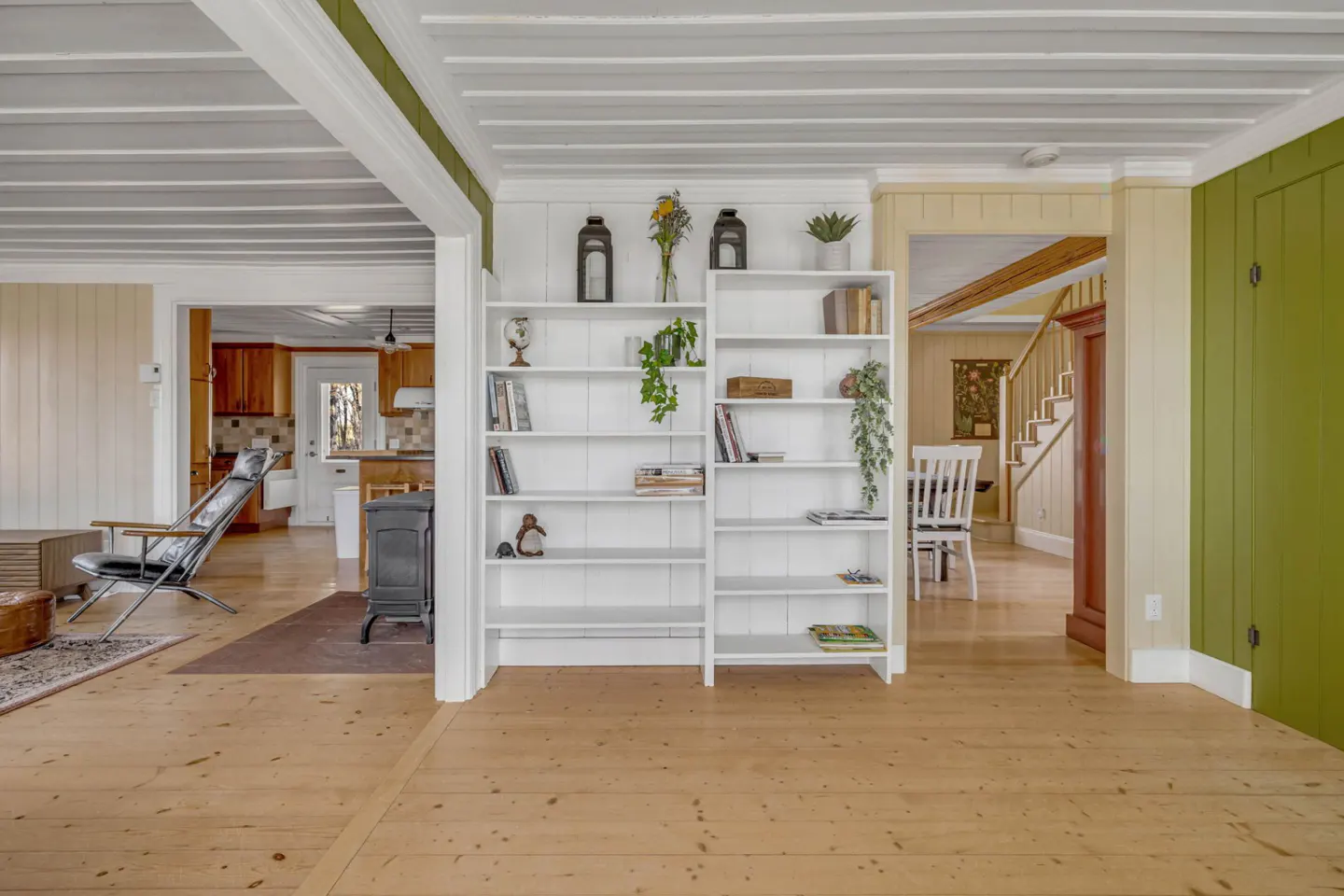 Interior view of a home with light wood floors, white bookshelves, and green accent walls. A kitchen and staircase are visible in the background.