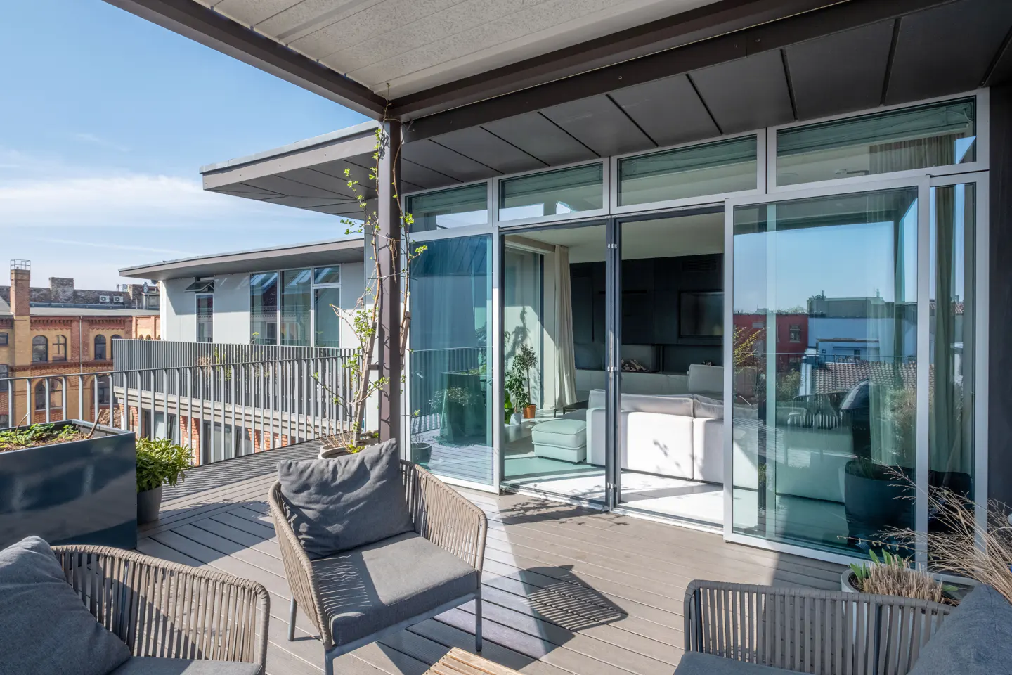 Modern rooftop terrace with gray wicker chairs, wooden decking, and glass sliding doors opening into a bright living room.