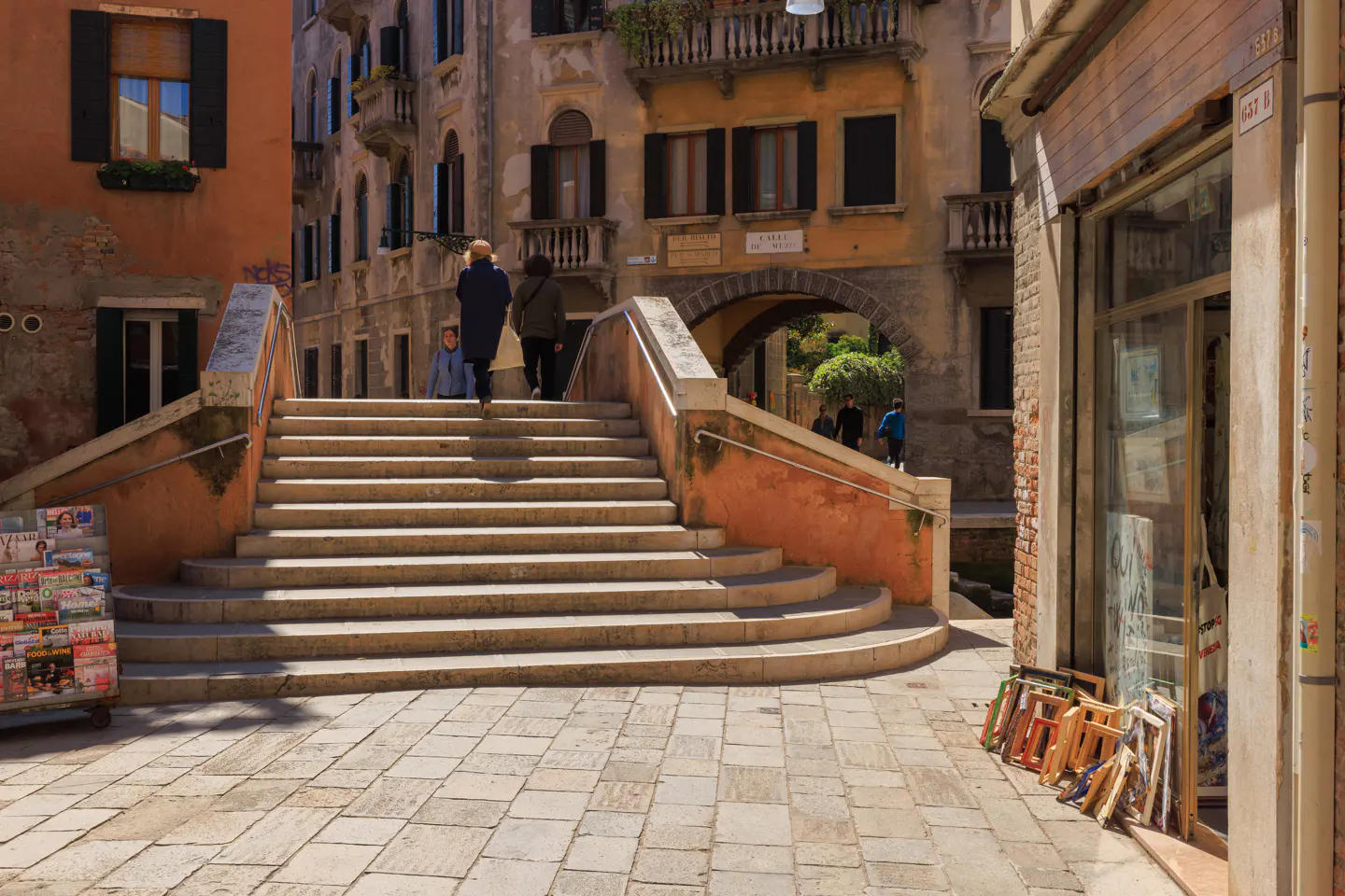 A sunny Venice street scene with people walking up stone steps over a canal. A magazine stand is on the left, and a shop is on the right.