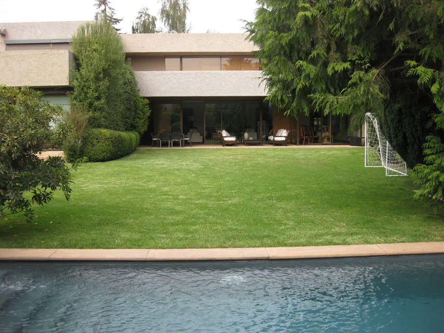 A modern house with a large green lawn, a swimming pool in the foreground, and a soccer goal in the background.